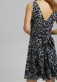 Ärmlös marinblå klänning med vitt blommönster, V-ringning på baksidan, figursydd midja med sammangjord tyg som knyts i en rosett på ryggen.