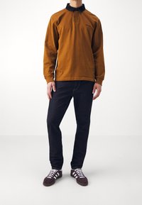 Hnědá polokošile s dlouhým rukávem a černým límcem, doplněná tmavě modrými džíny a hnědými teniskami s bílými pruhy. Jednoduchý design, bez vzorů.