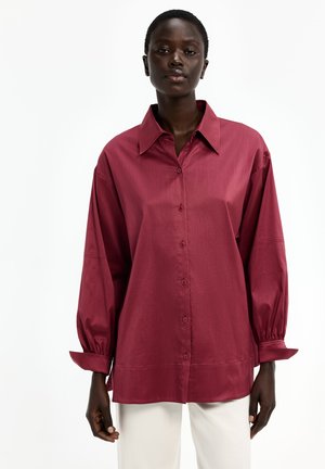 Frau trägt ein locker sitzendes, langärmliges dunkelrotes Hemd mit Knöpfen und weiße Hose, steht vor einem einfarbigen weißen Hintergrund.