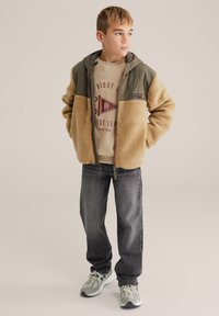 Barna és olívazöld polárbéléses kabát kapucnival, bézs színű pulóver grafikával, szürke farmernadrág és világos sportcipő.