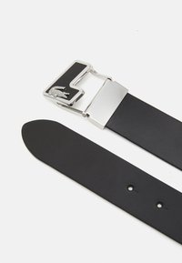 Cinto de couro preto com uma fivela metálica prateada e logotipo em relevo. A fita tem uma textura suave com múltiplos buracos de ajuste.