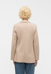 Personne aux cheveux blonds courts et bouclés, portant un blazer beige et un jean bleu, vue de dos sur un fond uni.