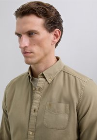 Mand med kort brunt hår iført en beige skjorte med knapper og lomme samt en hvid t-shirt, der ser til venstre mod en ensfarvet baggrund.