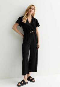 Jumpsuit noir à manches courtes avec décolleté en V et taille cintrée, mettant en valeur une ceinture ronde. Mannequin portant des sandales noires plates, sur un fond clair.