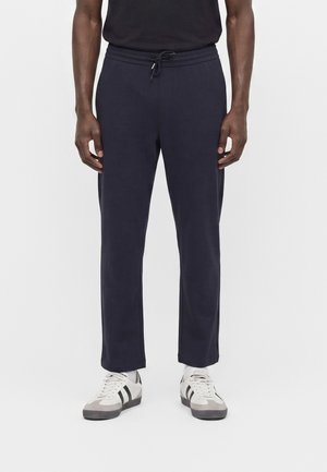 Mand iført marineblå bukser med snøre og hvide sneakers med sorte striber, stående mod hvid baggrund.