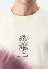 Svetăr crem cu un gradient violet în partea de jos, având un imprimeu floral negru și textul "marimekko". Material moale din bumbac.
