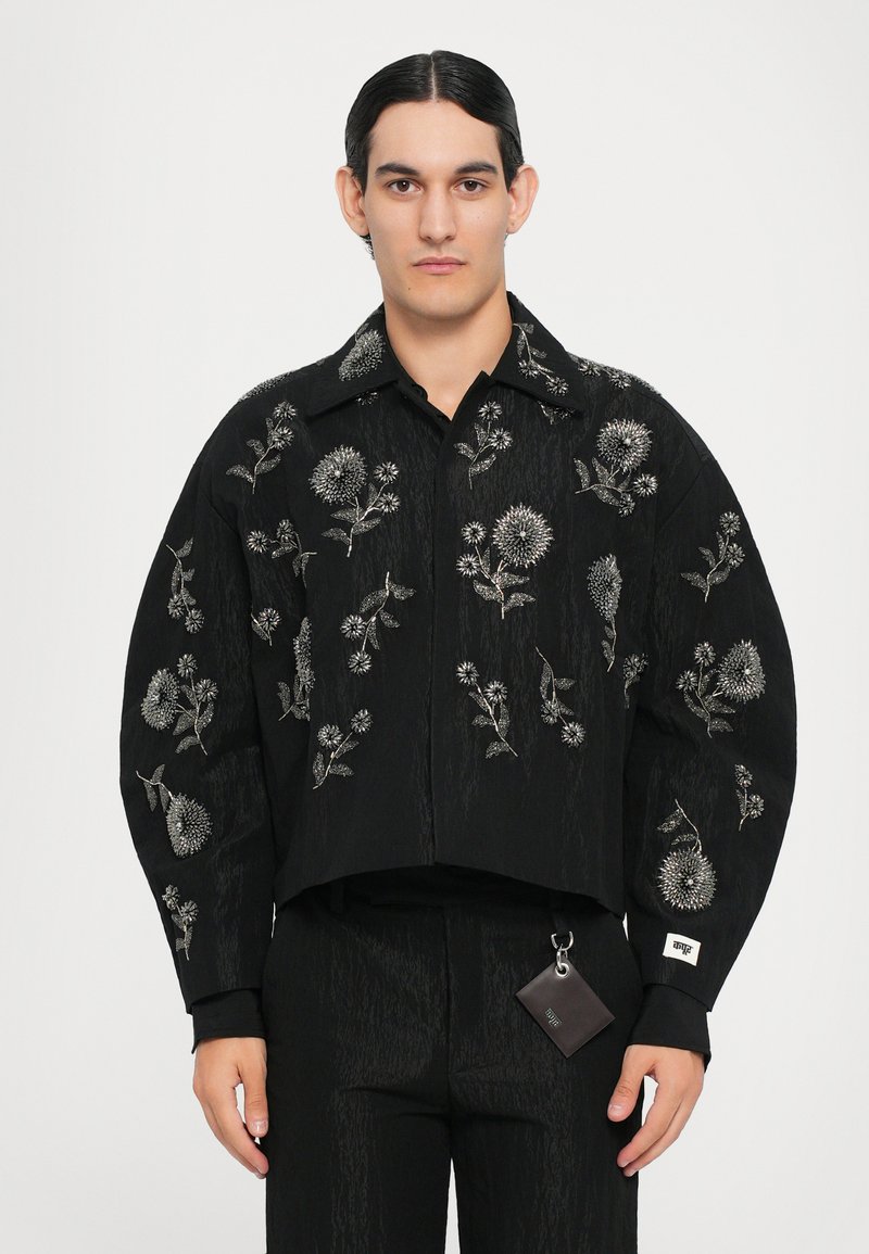 Schwarze, kurz geschnittene Jacke mit filigraner silberner Blumenstickerei, klassischem Kragen und langen Ärmeln. Inklusive eines kleinen Etiketts, das an der Taille befestigt ist.