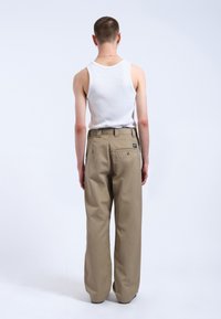 Lichtbeige broek met een wijde pijp en een ritsluiting, voorzien van riemlussen. Witte tanktop zichtbaar, van gladde en strakke stof.