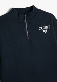 Sudadera azul marino con cuello alto y media cremallera. Presenta el texto "COURT" bordado en blanco y un gráfico de una raqueta de tenis en el pecho. Textura suave.