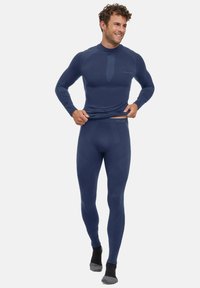 Man wearing fitted navy blue thermal top and leggings, pulling up shirt slightly, smiling, standing against plain background.