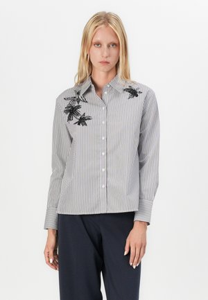 Νεαρή γυναίκα που φοράει πουκάμισο με κάθετες μαύρες και λευκές ρίγες και μαύρη κεντημένη λουλουδάτη διακόσμηση, καθώς και μπλε σκούρο παντελόνι, στέκεται μπροστά σε λευκό φόντο.