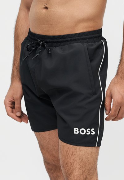 Homme portant un short de bain noir avec un passepoil blanc et le logo "BOSS" sur la jambe gauche, taille élastique avec cordon noir, longueur mi-cuisse.