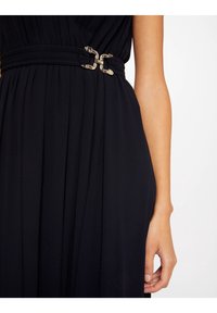 Svart klänning med plisserad design och midjebälte med silverfärgade spännen, som framhäver den släta tygtexturen och den skräddarsydda formen.