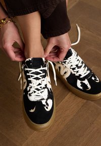 Zwart-wit gestreepte sneakers met een suede afwerking, voorzien van een rubberen zool en witte veters, die door handen worden geregen op een houten vloer.