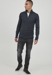 Marineblå zip-up genser i et mykt, strukturert stoff. Har høy krage og ribbede mansjetter, sammen med grå jeans og svarte sneakers.