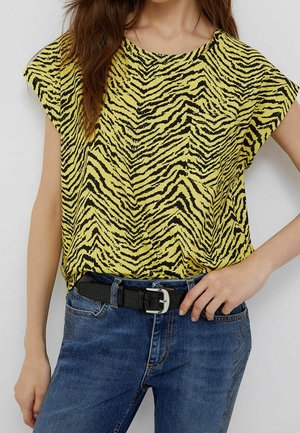 Kobieta w żółto-czarnym topie w paski zebry, włożonym do niebieskich dżinsów z czarnym paskiem, z jedną ręką na biodrze.