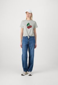Camiseta de rayas de manga corta con nudo en el frente, que presenta un gráfico de cereza. Combinada con jeans azul holgados y zapatillas blancas. Incluye una gorra blanca.