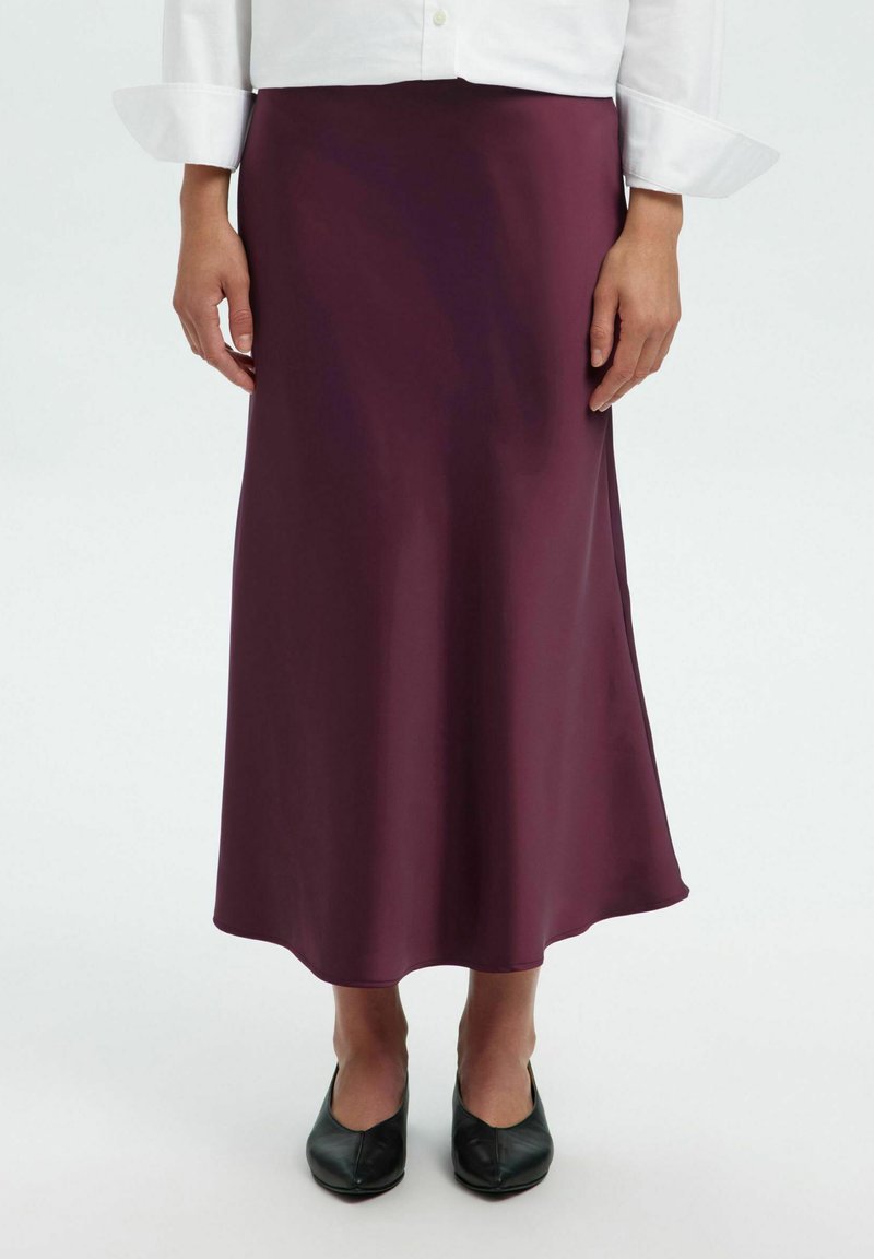 Gonna midi in satin bordeaux con una texture liscia e una silhouette fluida, abbinata a ballerine nere. L'orlo è leggermente curvo nella parte inferiore.