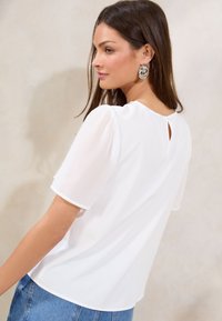Weiße Bluse mit kurzen, puffigen Ärmeln. Verfügt über einen Schlüssellochausschnitt im Rücken und eine glatte Textur. Kombiniert mit blauen Jeans. Sichtbare silberne Creolen.
