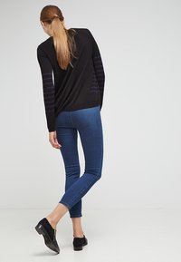 Sudadera negra con mangas a rayas moradas, combinada con jeans ajustados de color azul claro. El modelo lleva unos zapatos planos de ante negros. Diseño simple y moderno.