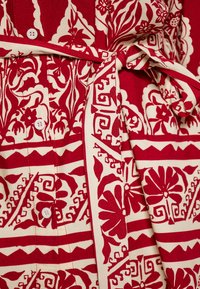 Kjolen i bomuldsstof har et rødt og cremefarvet blomstermønster med et bælte med snøring, knapper og intrikate designs langs kanterne.