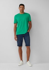 Zielony t-shirt z krótkim rękawem w połączeniu z ciemnymi szortami dżinsowymi o prostym kroju i podwiniętych nogawkach, uzupełniony białymi sneakersami.