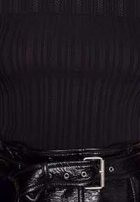 Gros plan sur un haut en maille côtelée noire rentrée dans un pantalon en cuir verni noir brillant avec une ceinture noire et une boucle argentée.