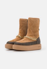 Beżowe zamszowe botki za kostkę z pikowanym brązowym wzorem, złotym łańcuszkiem na przodzie oraz masywną gumową podeszwą.