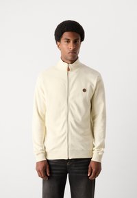Cremefarvet zip-up jakke med høj krave, ribbede ærmer og et lille logo på venstre bryst. Blødt, struktureret stof, tætsiddende design.