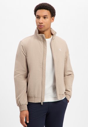 Jonge man draagt een beige jas met rits over een wit overhemd en donkere broek, kijkt zijwaarts tegen een effen achtergrond.