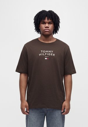 Jeune homme aux cheveux bouclés portant un t-shirt Tommy Hilfiger marron foncé et un jean bleu, debout devant un fond clair uni.