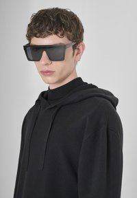 Felpa nera oversize con finitura testurizzata, indossata sopra un dolcevita nero, abbinata a occhiali da sole neri angolari con lenti scure.