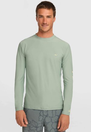 Man met lichtgroen lang mouwen hemd en grijze korte broek met bloemenpatroon, staand tegen een effen lichte achtergrond.