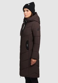 Vrouw in een lange, donkergroene, gewatteerde winterjas met capuchon en een zwarte gebreide muts tegen een effen lichtgrijze achtergrond.