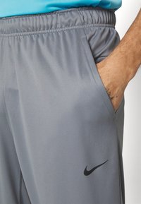 Manuell hand som vilar i fickan på grå Nike träningsoverallsbyxor med elastisk midja och svart Nike swoosh-logotyp.