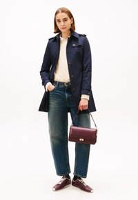 Marineblå trenchcoat med knappedetaljer og skulderklaffer, kombinert med en kremfarget kabelstrikket genser, blå jeans og en burgunder håndveske.