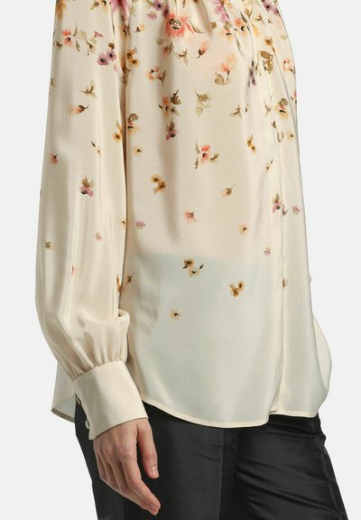 Woman wearing a long-sleeve, cream blouse with small floral print paired with black pants, hand relaxed by her side.