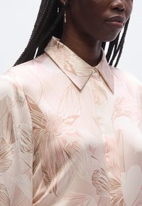 Μεταξωτό μπλουζάκι με ανοιχτό ροζ βάση που διαθέτει μεγάλα ανθικά σχέδια σε σκοτεινότερο ροζ και εκρού. Σχεδιασμός με γιακά και λεπτομέρειες με κουμπιά μπροστά.