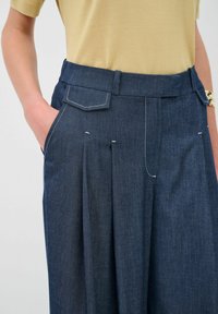 Pantaloni in denim a vita alta, con pieghe frontali, tasche posteriori e cuciture bianche a contrasto. Abbinati a un top giallo a maniche corte.