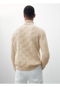 Beiger Strickpullover mit hohem Kragen und Wellenmuster, mit strukturiertem Rippstrick und tailliertem Schnitt, getragen über weißer Hose.