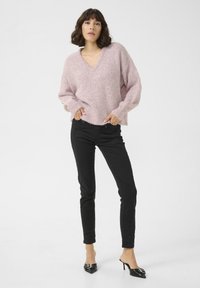 Pulover tricotat de culoare roz deschis, cu decolteu în V și tiv cu nervuri, asociat cu pantaloni negri ajustați și mules negre cu toc, decorate cu detalii din hardware.