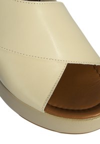 Beige leather sandal with an open toe, crisscross design, and brown interior padding. Features a textured sole and minimal stitching.