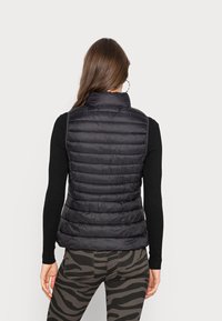 Schwarze, puffy Weste mit hohem Kragen, gestepptem Muster und Seitentaschen. Getragen über einem schwarzen Langarmtop mit enganliegendem Ärmel.