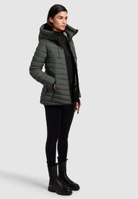 Gepolsterte dunkelgrüne Jacke mit Kapuze, Reißverschluss, Seitentaschen und gerippten Bündchen, kombiniert mit schwarzen Leggings und klobigen schwarzen Stiefeln.