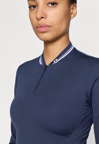 Top atletico blu navy con chiusura a zip, caratterizzato da un colletto a coste con strisce bianche e pannelli laterali testurizzati per un tocco di dettaglio in più.