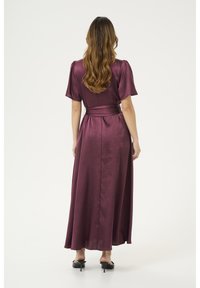 Long, burgundy dress with short puff sleeves, a waist tie, and a flowing skirt. Fabric has a shiny texture, with a back slit for movement.
