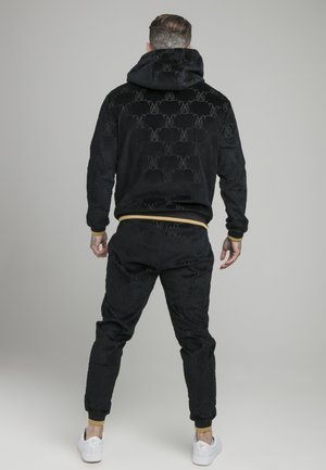 Mand set bagfra iført sort mønstret hættetrøje og matchende bukser med tan manchetter samt hvide sneakers mod en ensfarvet grå baggrund.