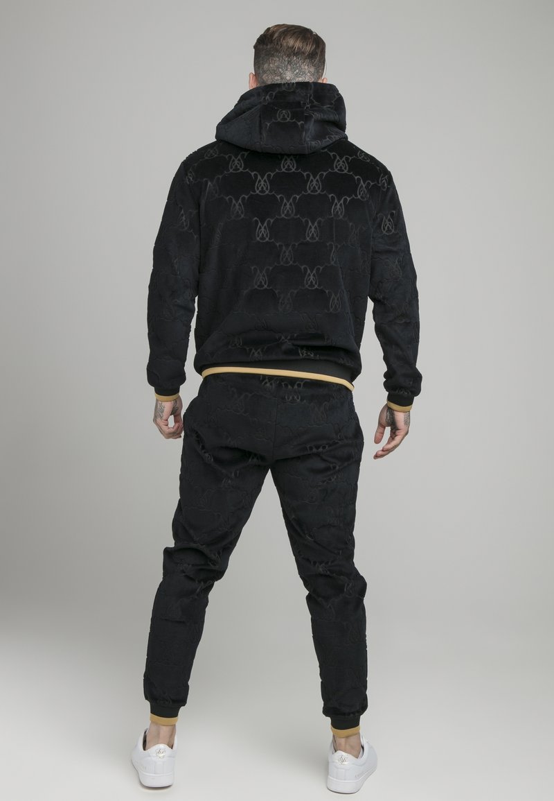 Homme vu de dos portant un sweat à capuche noir à motifs et un pantalon assorti avec des poignets couleur beige, ainsi que des baskets blanches sur un fond gris uni.