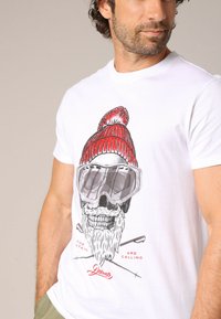 T-shirt blanche avec un design graphique représentant un crâne portant un bonnet rouge et des lunettes de ski, accompagné de bâtons de ski croisés et d'un texte en dessous.