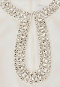 Biely materiál s prepracovaným perlovým golierom z rôznych jasných kryštálov rôznych tvarov a veľkostí. Zapínanie na zips na zadnej strane.